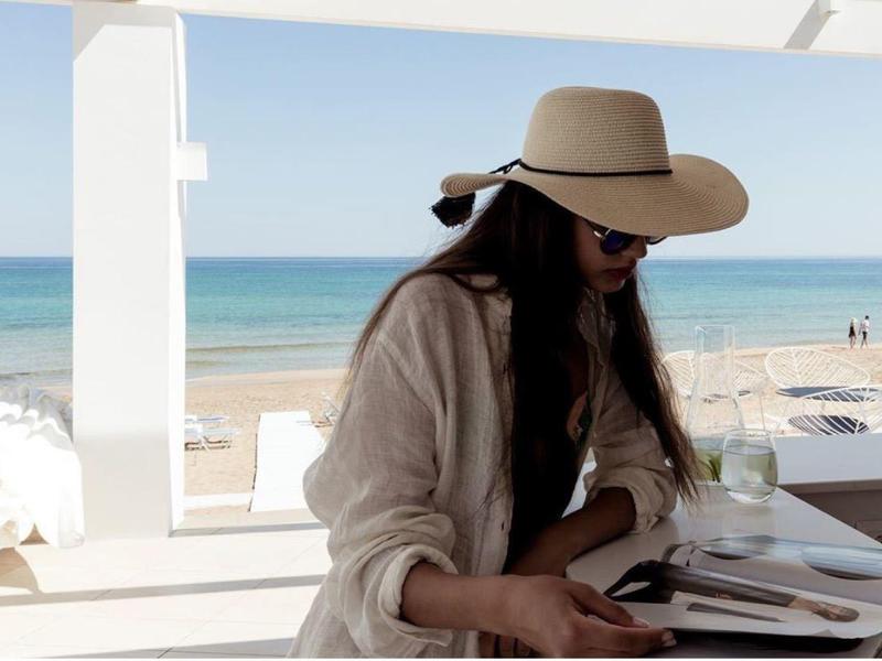 Žena s kloboukem sedí u stolu u moře a čte knihu na pláži.