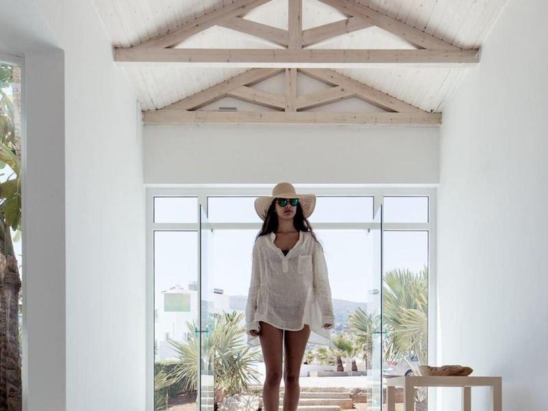 Žena s kloboukem v bílém tričku stojí v světlé místnosti s dřevěnými trámy a výhledem na přírodu.