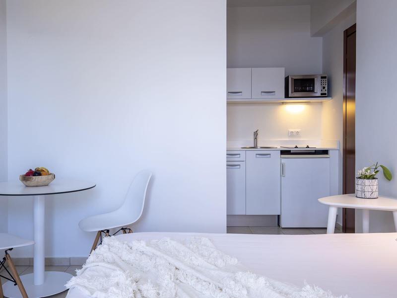 Minimalisticky zařízený hotelový pokoj se stolem, židlí a kompaktní kuchyňkou.