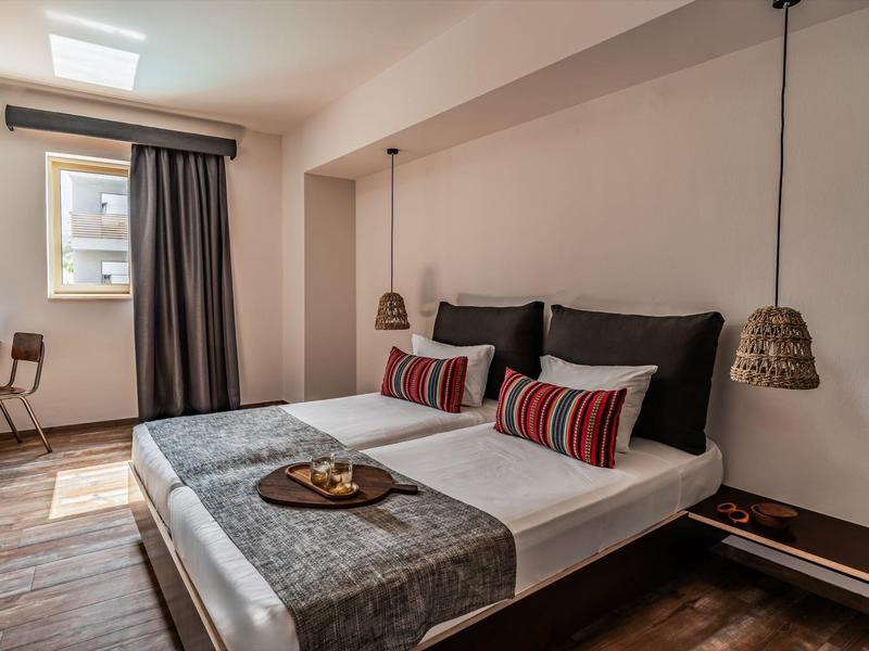 Moderní hotelový pokoj s manželskou postelí, dekorativními polštáři, psacím stolem a oknem s závěsem.