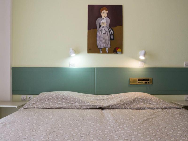 Dvoulůžko s šedým povlečením před zelenou zdí s dětským obrazem a dvěma čtecími lampami.