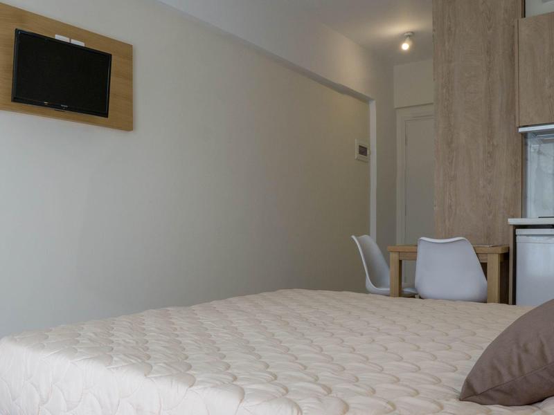 Jednoduchý hotelový pokoj s postelí, židlemi, stolem a televizí na stěně.