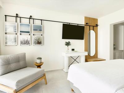 Helles Schlafzimmer mit weißem Bett, grauem Sessel, TV an der Wand und Holzspiegel mit Ablage.