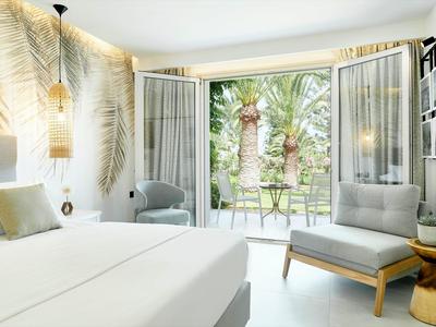 Helles Schlafzimmer mit weißem Bett, grauem Sessel, Palmenmuster an der Wand und Gartenblick.