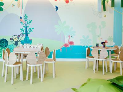 Buntes Kinderzimmer mit weißen Stühlen, Tischen und einer Wand mit fröhlichen Tier- und Naturmotiven.