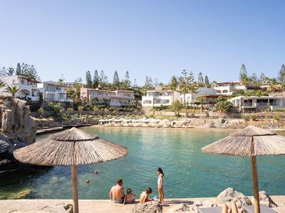 Strandbereich mit Strohsonnenschirmen, Menschen am Wasser und Reihen von Ferienhäusern im Hintergrund.