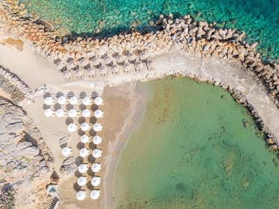 Luftaufnahme eines Strandes mit weißen Sonnenschirmen, Felsen und klarem türkisfarbenem Wasser.
