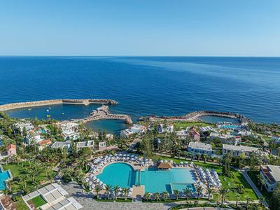 Blick auf eine Küstenanlage mit Hotel, zwei Pools, grünen Flächen und einem Hafen im blauen Meer.