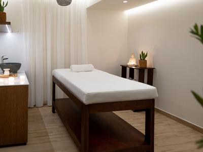 Heller Massageraum mit Holzliege, weißen Wänden, grünen Pflanzen und sanfter Beleuchtung.