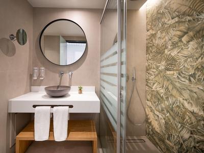 Nowoczesna łazienka z okrągłym lustrem, umywalką na białej szafce i kabiną prysznicową z roślinnym wzorem na ścianie.