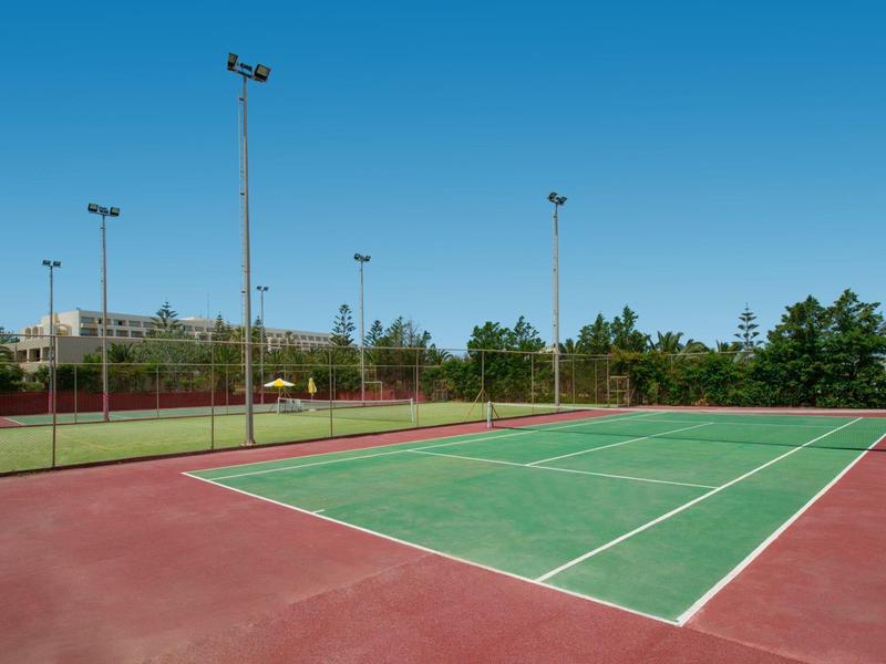 Freier Tennisplatz mit grünem Spielfeld, roten Außenflächen und blauem Himmel im Hintergrund.