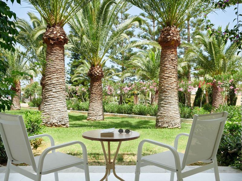 Blick von Terrasse mit zwei Stühlen und kleinem Tisch auf grünen Garten mit Palmen.