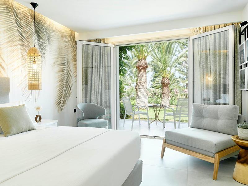 Helles Schlafzimmer mit weißem Bett, grauem Sessel, Palmenmuster an der Wand und Gartenblick.