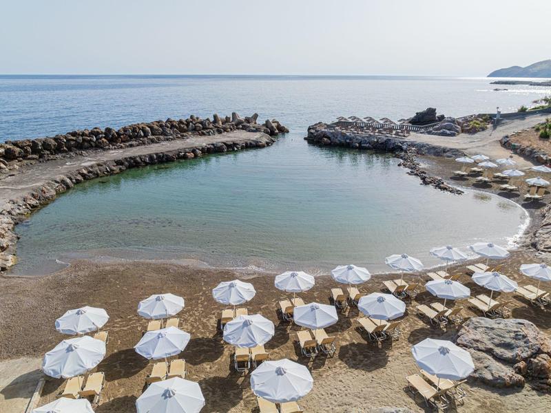 Kleiner Strand mit klarem Wasser, vielen weißen Sonnenschirmen und felsigen Uferabschnitten.