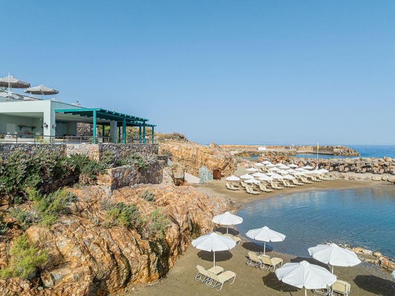Kleiner Strand mit Felsen, weißen Sonnenschirmen und Liegen, angrenzend an modernes Gebäude.
