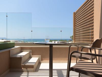 Balkon mit Stuhl, Tisch und Glasgeländer, Blick auf Meer und Strand bei klarem Himmel.