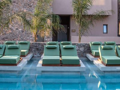 Plusieurs chaises longues vertes sur des plateformes dans une piscine avec un mur de pierre et des palmiers en arrière-plan.