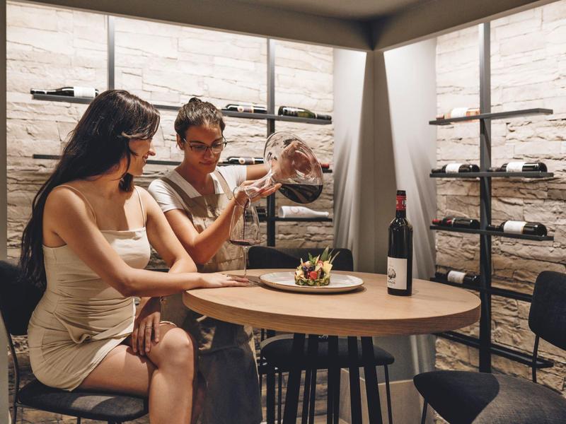 Dwie kobiety siedzą w piwnicy z winem, oglądając butelkę wina przy okrągłym stole.