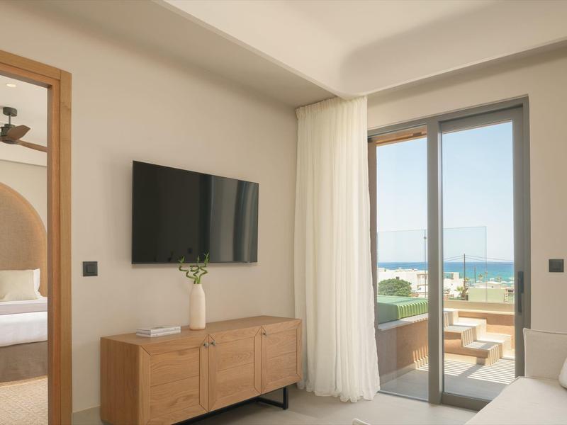 Nowoczesny pokój hotelowy z telewizorem, drewnianymi meblami i widokiem na taras i morze.