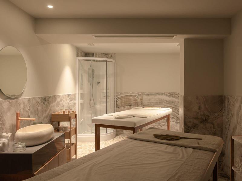 Jasne pomieszczenie spa z łóżkami do masażu, okrągłym lustrem i kabiną prysznicową z marmurowymi ścianami.