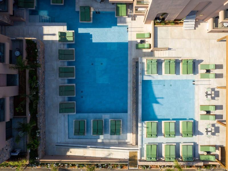 Widok z lotu ptaka na prostokątny basen z leżakami otoczony budynkami.