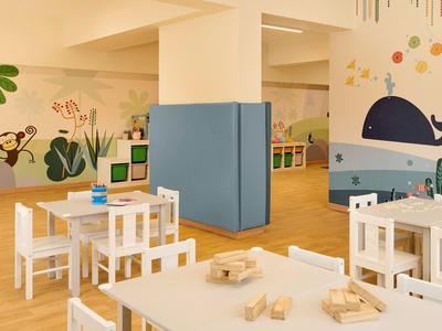 Heller Kindergartenraum mit Holzmöbeln, blauer Raumecke und bunten Tierwandbildern.