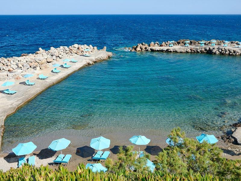 Eau claire entourée de rochers avec des parasols bleus et des chaises longues sur une petite plage.
