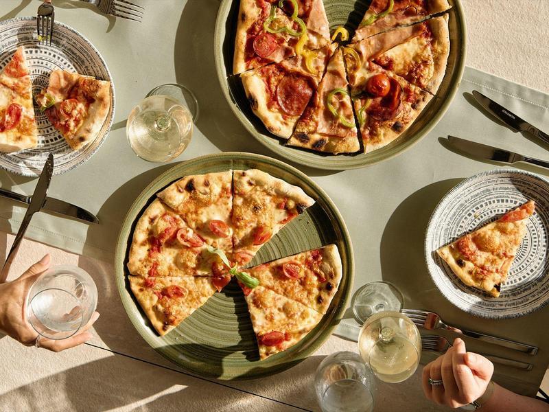 Deux personnes dégustent une pizza et du vin blanc à une table ensoleillée en plein air.