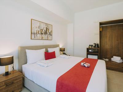 Nowocześnie urządzony pokój hotelowy z łóżkiem podwójnym, czerwonymi akcentami i otwartą szafą.