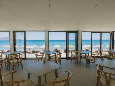 Widok na hotelową jadalnię z drewnianymi stołami i krzesłami przed dużymi oknami z widokiem na plażę.