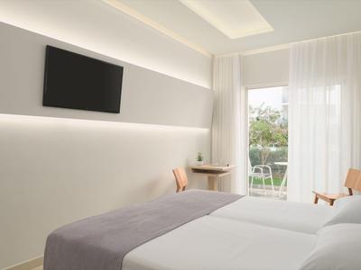 Jasny pokój hotelowy z dwoma łóżkami i balkonem z widokiem na zieleń.