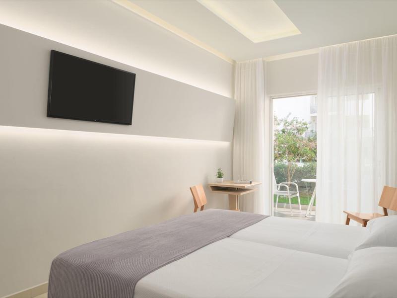 Habitación de hotel moderna con dos camas individuales, televisor en la pared y balcón con vista a la vegetación.