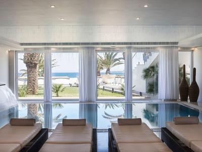 Helles Wellnessbad mit Pool, Liegen und großen Fenstern zum Garten mit Palmen und Meerblick.