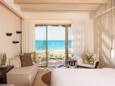 Helles Hotelzimmer mit großem Bett, Sofa, TV, Balkon und Meerblick bei Sonnenschein.