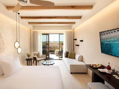 Helles Schlafzimmer mit Holzdeckenbalken, großem Bett, Sofa, Fernseher und Blick auf Terrasse und Landschaft.