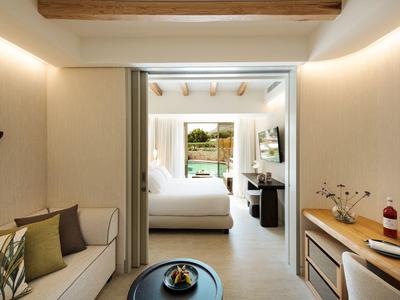 Hell eingerichtetes Zimmer mit Holzbalken, gemütlichem Bett, Sofa, Tisch und Blick auf Pool draußen.