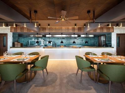 Modernes Restaurant mit grünen Stühlen, Holztischen und offenem Kochbereich mit Fliesenwand.
