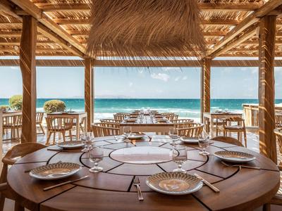 Offenes Strandrestaurant mit runden Holztischen, gedeckten Tellern und Blick auf das Meer.