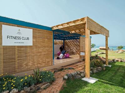 Modernes Fitnessclubhaus aus Holz mit Sitzbereich, umgeben von grünem Rasen und Meer im Hintergrund.