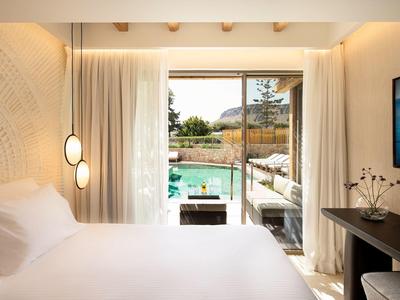 Helles Zimmer mit großem Bett, Vorhängen und Blick auf einen Pool und Garten durch Glasfenster.