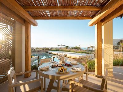 Terrasse mit rundem Holztisch, gedecktem Essen, Stühlen und Blick auf Wasser und Häuser im Hintergrund.