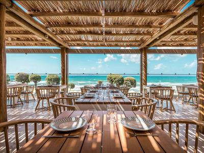 Restaurace pod širým nebem s dřevěnými stoly s výhledem na pláž a moře pod jasnou oblohou.