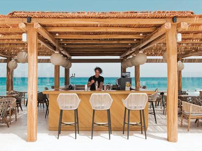 Plážový bar s dřevěnou konstrukcí, stoličkami a výhledem na modré moře v pozadí.