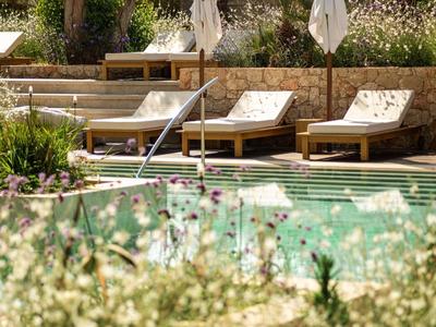 Una piscina con lettini in legno e un muro di pietra, circondata da piante verdi e fiori.