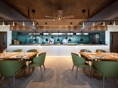 Interno di ristorante moderno con sedie verdi, tavoli apparecchiati e cucina a vista.