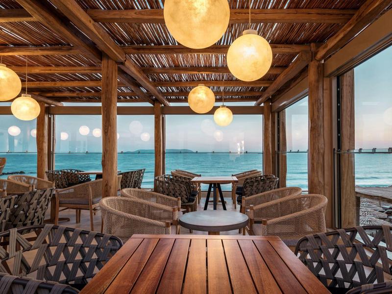 Accogliente bar sulla spiaggia con sedie rotonde, tavoli in legno e vista sul mare al tramonto.