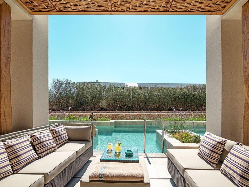 Accogliente zona salotto con cuscini che si affaccia su una piscina e un giardino sotto un cielo limpido.