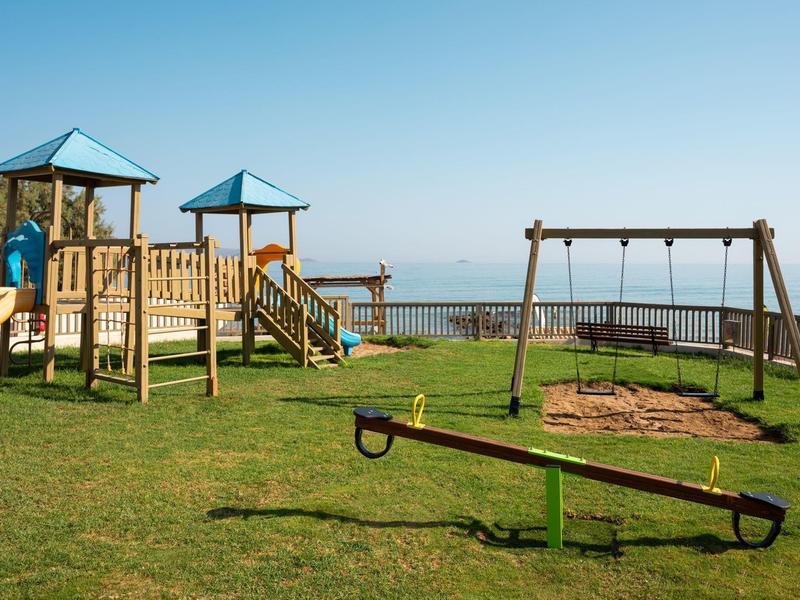 Parco giochi per bambini con scivolo, altalene e altalena a dondolo su prato con vista mare.