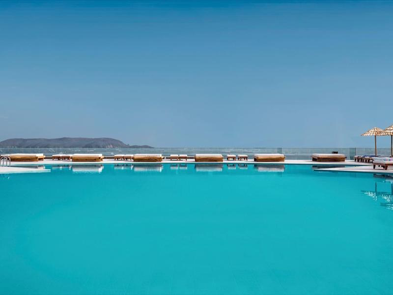 Grande piscina turchese con ombrelloni e lettini vicino al mare sotto un cielo sereno.