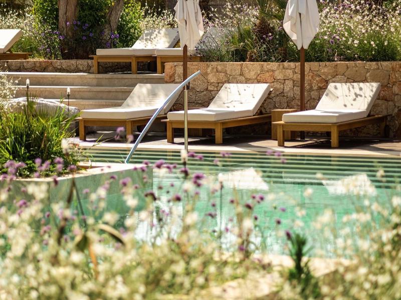 Una piscina con lettini in legno e un muro di pietra, circondata da piante verdi e fiori.